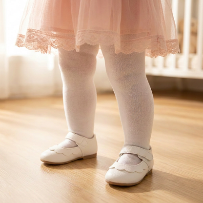 Toddler girl wearing white scalloped Mary Jane shoes with white tights and a pink lace tulle dress, standing on a hardwood floor in a bright room.