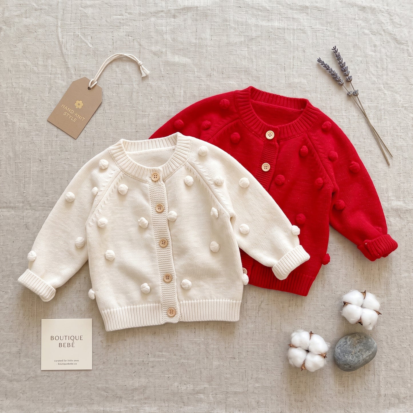 Flat lay showing both the red and cream colors of the baby girls cotton knit pom-pom cardigan sweaters.