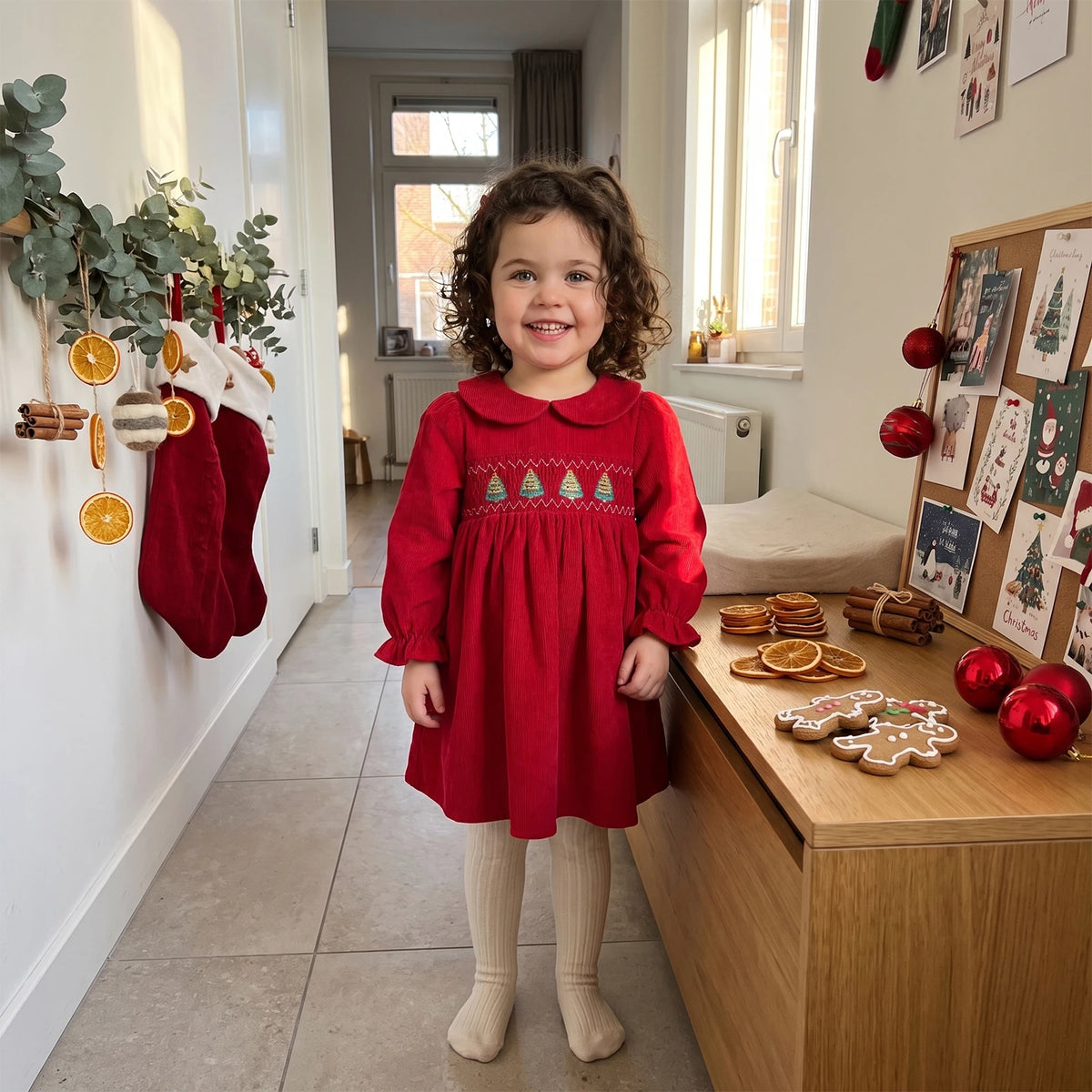 Young girl standing in a room wearing a red long-sleeve smocked corduroy Christmas dress with cream tights.