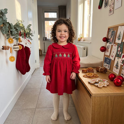 Young girl standing in a room wearing a red long-sleeve smocked corduroy Christmas dress with cream tights.