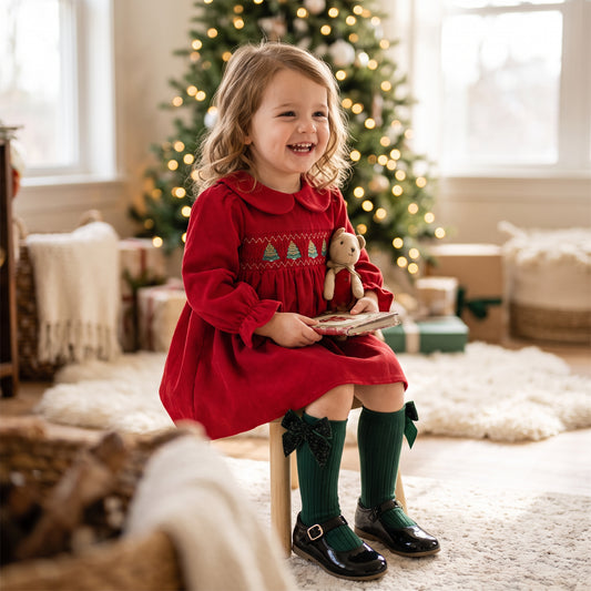 Toddler girl sitting on a stool wearing a red corduroy Christmas dress with green knee socks in front of a holiday tree.