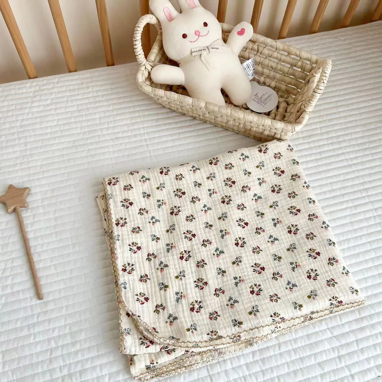 Red ditsy floral print cotton muslin receiving blanket softly draped in a baby crib with a woven basket and bunny plush.