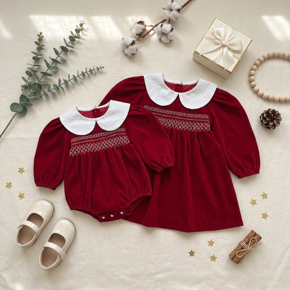 A flat lay presentation of a matching red smocked baby romper and toddler dress, styled with baby shoes, a gift box, and winter greenery.