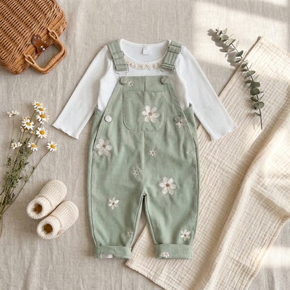 Styled flat lay featuring sage green Kidsely daisy overalls layered over a white long-sleeve top, paired with baby shoes and dried florals.