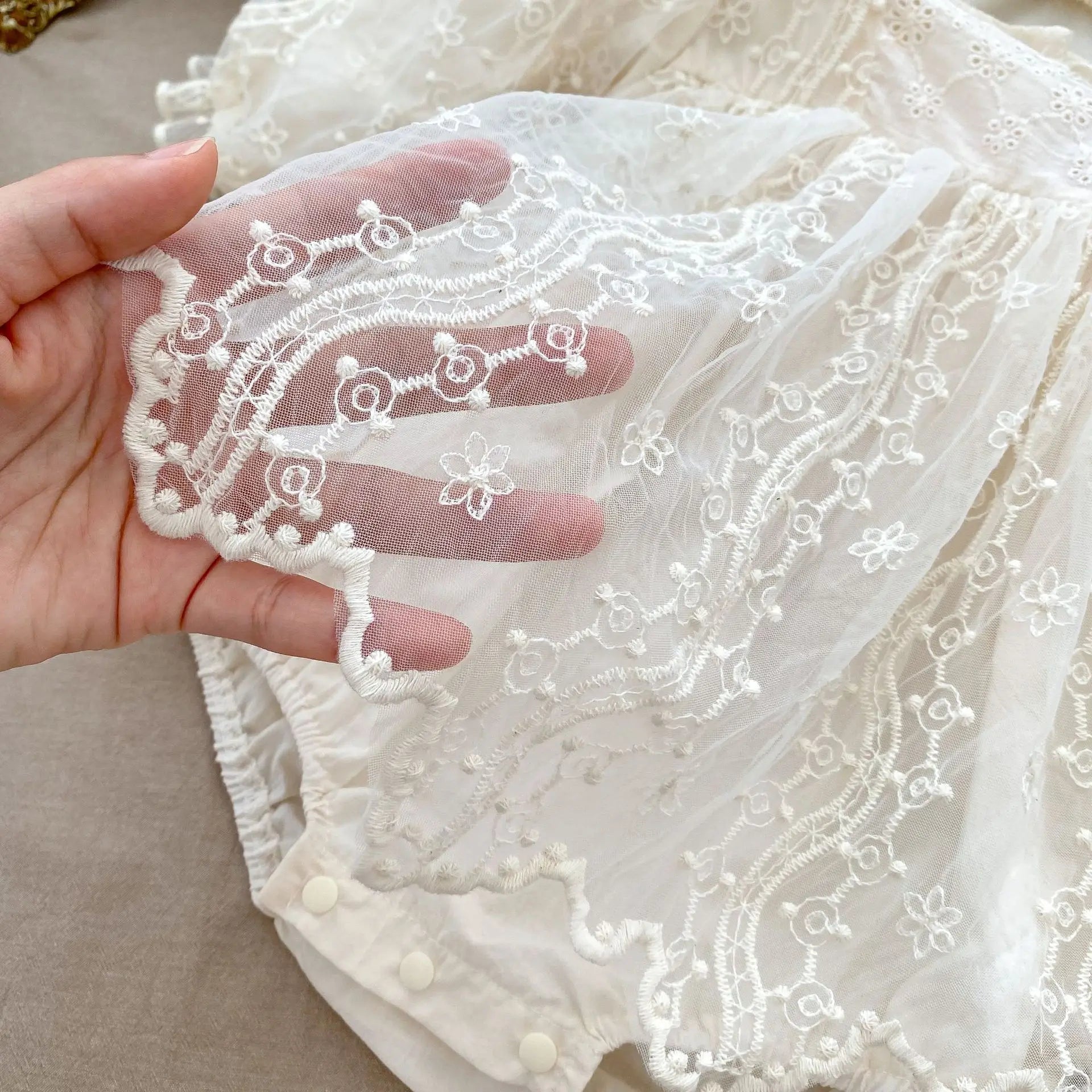 Hand lifting the sheer mesh lace overlay of a baby girl romper to show the scalloped trim and floral embroidery.