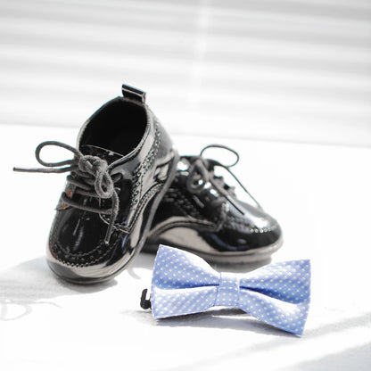 Close-up of a pair of high-gloss black Kidsely toddler dress shoes arranged on a bright surface next to a light blue patterned baby bowtie.