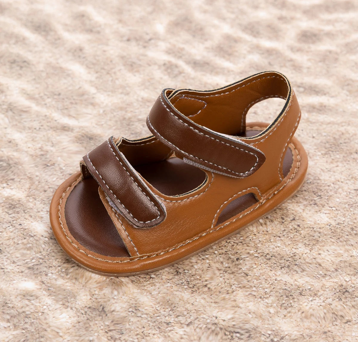 A single brown vegan leather baby sandal sitting on a sandy background.