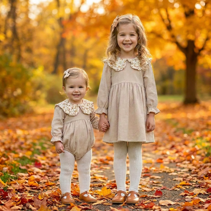 Two sisters holding hands outdoors in fall leaves, wearing matching beige ribbed corduroy outfits featuring a baby romper and toddler dress.