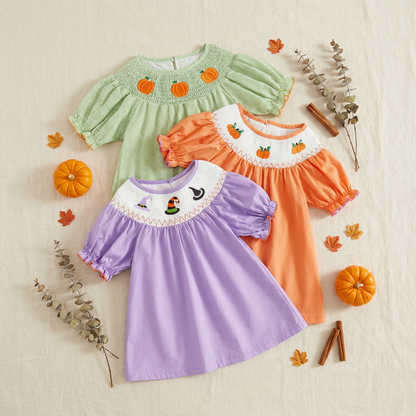 Flat lay display of three solid-colored smocked fall dresses in light green, purple, and orange, surrounded by autumn decor.