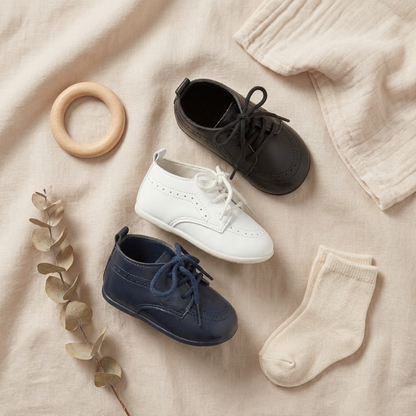 Modern aesthetic flat lay displaying Kidsely smooth white, black, and navy baby oxfords styled next to a wooden teething ring and cream-colored knit socks.