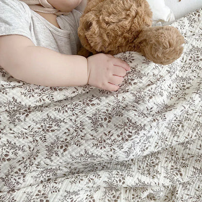 Close up of a baby's hand resting on a brown teddy bear while covered by a soft taupe leaf print cotton muslin blanket.