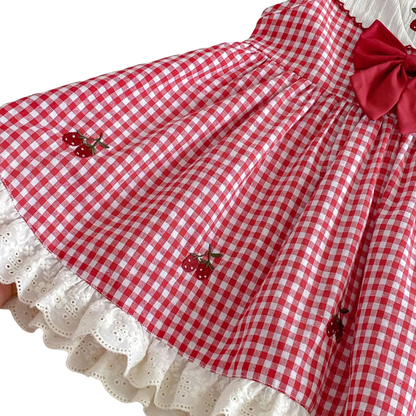 Close-up detail of high-quality red strawberry embroidery on a red and white gingham toddler dress; features a white eyelet lace ruffle hem and textured fabric.