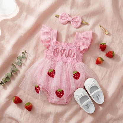 Flat lay styling of the pink strawberry first birthday romper outfit, headband, white shoes, and scattered fresh strawberries