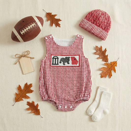 Styled flat lay of a red seersucker gingham infant gameday romper surrounded by fall football tailgate accessories including a mini football, red knit baby beanie, white socks, and autumn leaves on a cream surface.