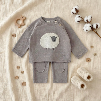 Styled aesthetic flat lay of the grey knit sheep baby outfit arranged on a textured blanket with cotton sprigs and knit baby booties.