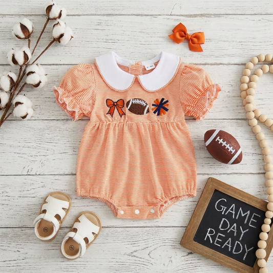 Styled flat lay of an orange striped baby game day romper surrounded by white sandals, an orange bow, a mini football, and a small chalkboard sign reading Game Day Ready.