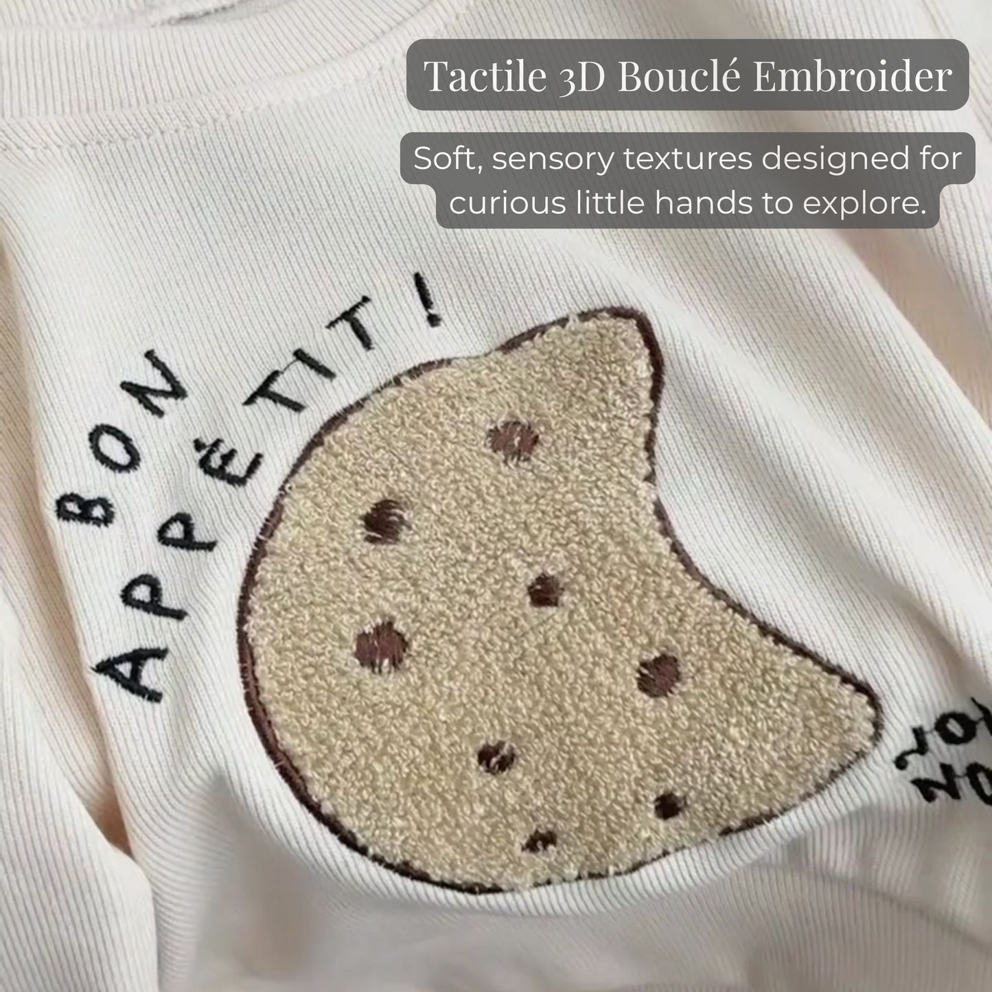 A close-up of the tactile 3D boucle cookie embroidery and "bon appétit!" lettering on the ribbed cream baby sweatshirt, highlighting the sensory texture.