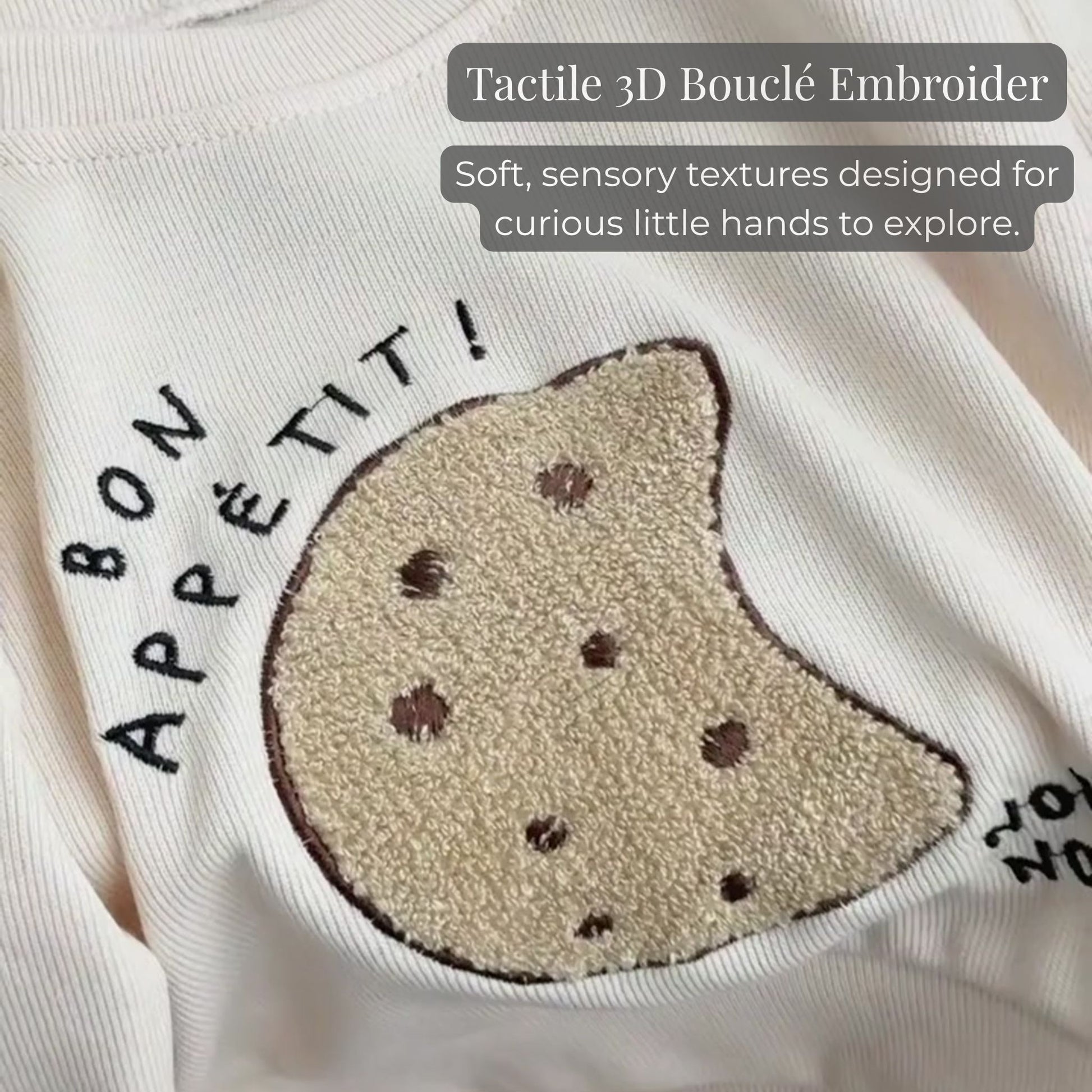 A close-up of the tactile 3D boucle cookie embroidery and "bon appétit!" lettering on the ribbed cream baby sweatshirt, highlighting the sensory texture.