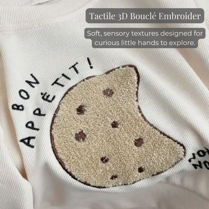A close-up of the tactile 3D boucle cookie embroidery and "bon appétit!" lettering on the ribbed cream baby sweatshirt, highlighting the sensory texture.