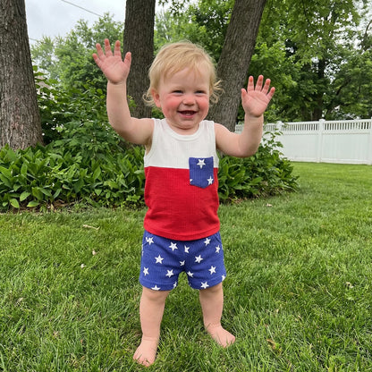 Toddler boy smiling and playing outside in the grass wearing a 2-piece red, white, and blue star-print summer outfit.
