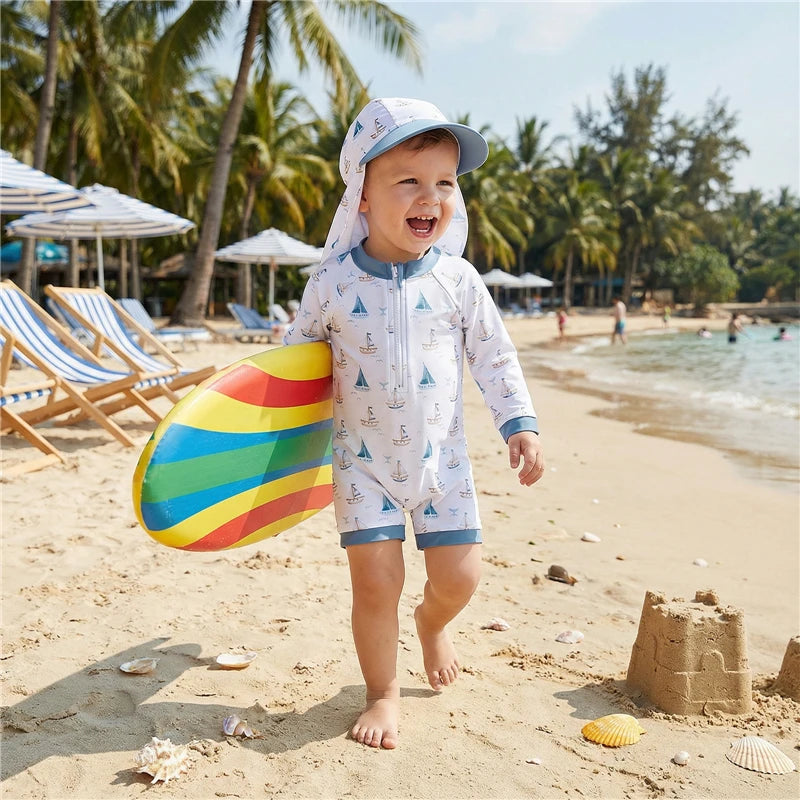 Child in a long sleeve nautical sailboat rash guard swimsuit and hat on a beach with a colorful sandcastle and palm trees in the background.