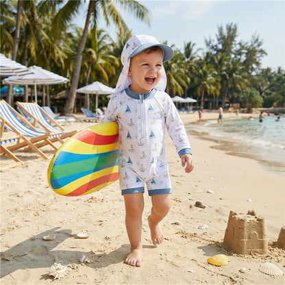 Child in a long sleeve nautical sailboat rash guard swimsuit and hat on a beach with a colorful sandcastle and palm trees in the background.