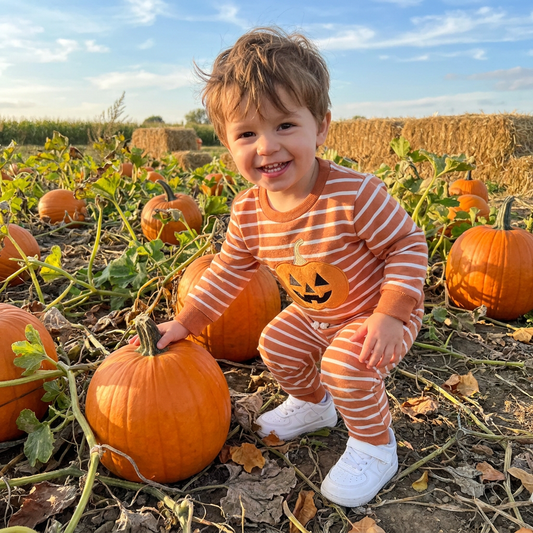 A smiling toddler boy wearing the orange and white striped Halloween pumpkin sweatshirt and jogger set while playing outdoors in a fall pumpkin patch.