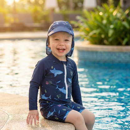 Child wearing a navy blue long sleeve shark swimsuit with sun hat sitting by a pool.