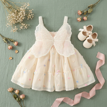 Creative flat lay presentation of the butterfly wing princess dress on a soft green background, styled alongside matching cream baby shoes, a pink ribbon, and dried flowers.