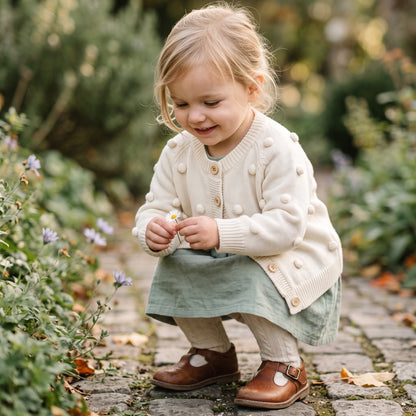 Toddler girl smiling outdoors wearing the cozy cream cotton knit pom-pom cardigan sweater over a dress.