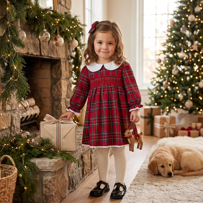 Little girl wearing a long sleeve red plaid smocked Christmas dress standing by a holiday fireplace