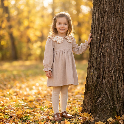 A little girl standing by a tree in an autumn setting, wearing a beige long-sleeve corduroy dress with a floral collar and cream tights.