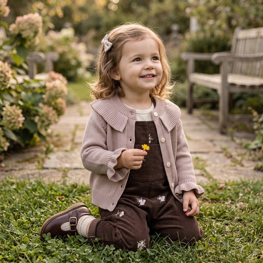 A young toddler girl sitting on grass in a garden, wearing a taupe cardigan over a white top and dark brown corduroy overalls with embroidered details.