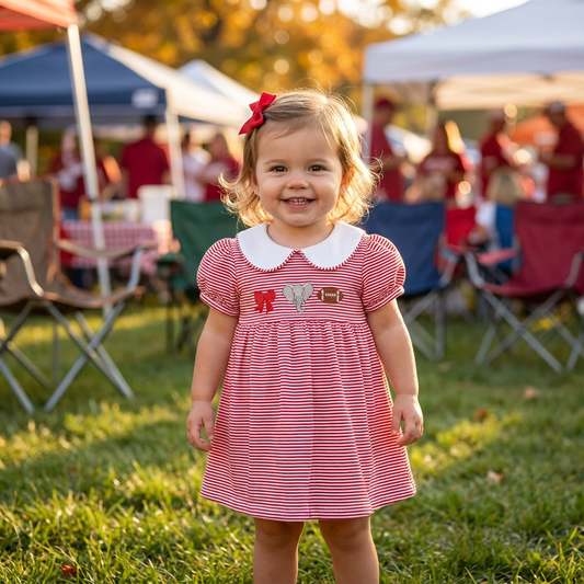 A smiling toddler girl wearing the crimson red striped game day dress with elephant embroidery, standing outdoors on the grass at a football tailgate.