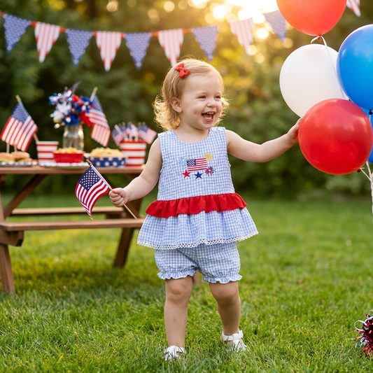 Smiling toddler girl wearing a blue gingham 4th of July outfit with red ruffles, holding an American flag at a summer picnic.