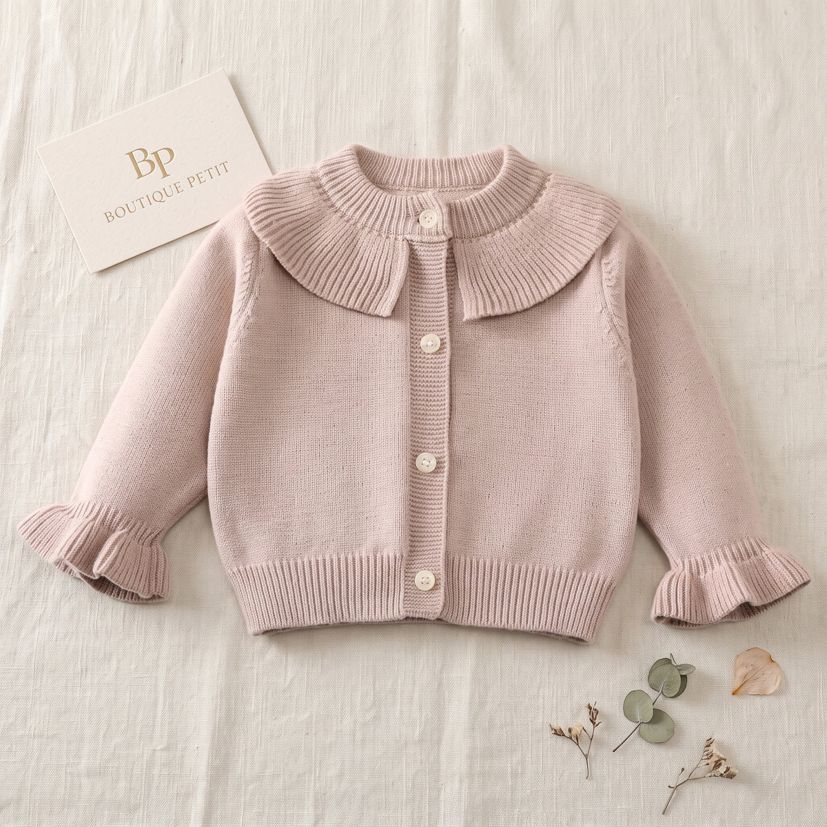 Toddler girl sitting and playing, wearing a soft taupe ruffle collar knit cardigan sweater over a floral dress.