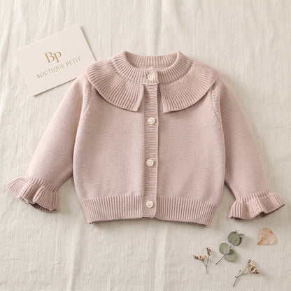 Toddler girl sitting and playing, wearing a soft taupe ruffle collar knit cardigan sweater over a floral dress.