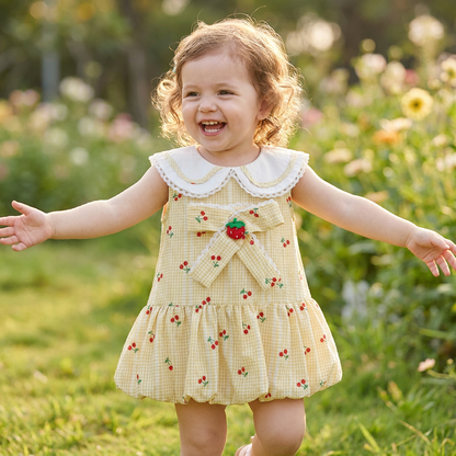 A smiling toddler girl playing outdoors with arms outstretched, wearing the yellow cherry gingham bubble dress.