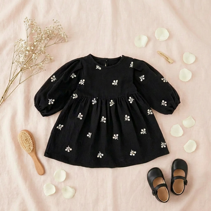 Flat lay of a toddler girls black floral embroidered puff sleeve dress on a soft pink background, styled with black Mary Jane shoes, a wooden hairbrush, a gold hair clip, and white flower petals.