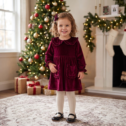 Toddler girl standing in front of a Christmas tree wearing a wine red velvet A-line holiday dress with white tights.