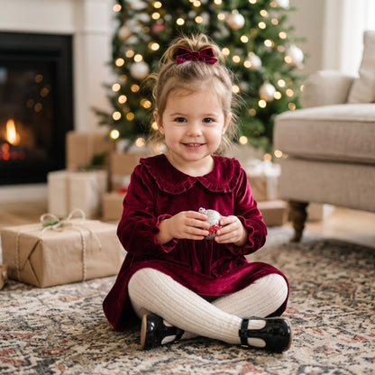 Toddler girl sitting near wrapped Christmas gifts wearing a wine red long sleeve velvet holiday dress and matching hair bow.