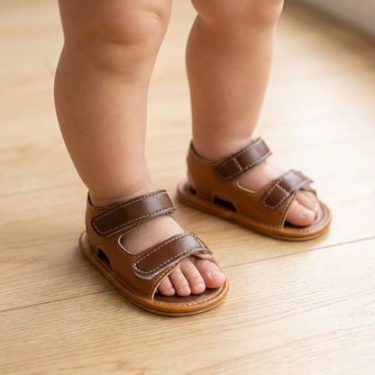 Close-up of a baby's legs standing on a light wood floor wearing brown double-strap vegan leather sandals.