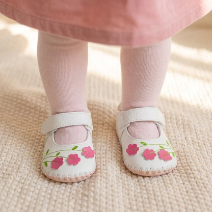 A detailed shot of a toddler's legs wearing pink tights and the white floral Mary Jane shoes, standing on a neutral surface, demonstrating fit and walking stability.