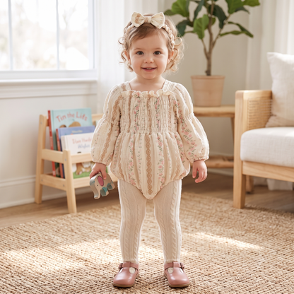 A smiling toddler girl standing in a nursery wearing a vintage floral cotton gauze bubble romper layered with white tights and a matching headband.