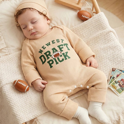 A baby sleeping peacefully on a cozy white blanket wearing a beige "Newest Draft Pick" football romper and hat, with a small football prop nearby.