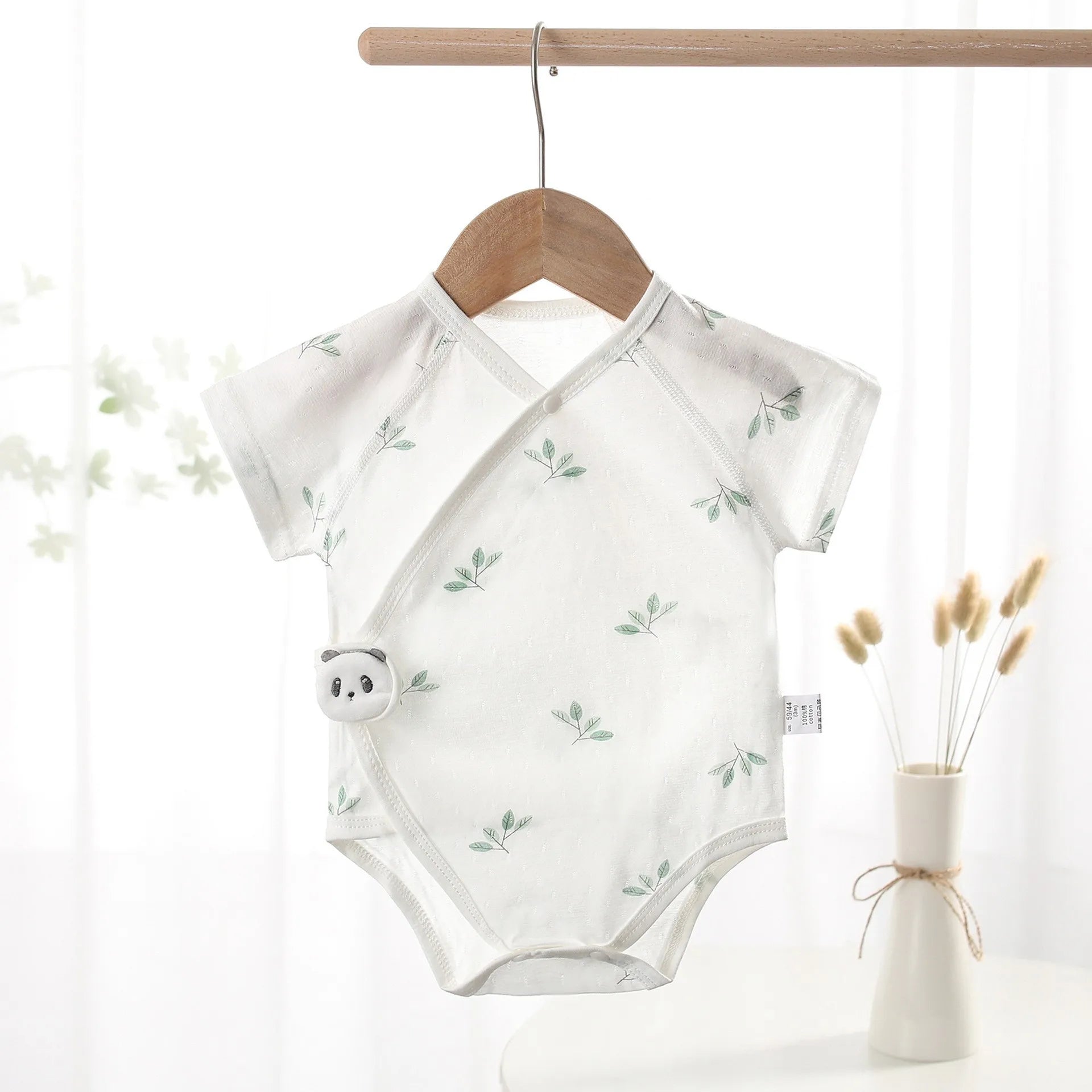 Unisex white botanical leaf print baby bodysuit featuring a plush panda detail, hanging on a wooden hanger next to a vase with dried pampas grass.