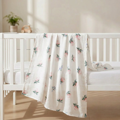 A large bamboo cotton muslin baby swaddle blanket featuring a watercolor pink rose floral print, draped over a white nursery crib.