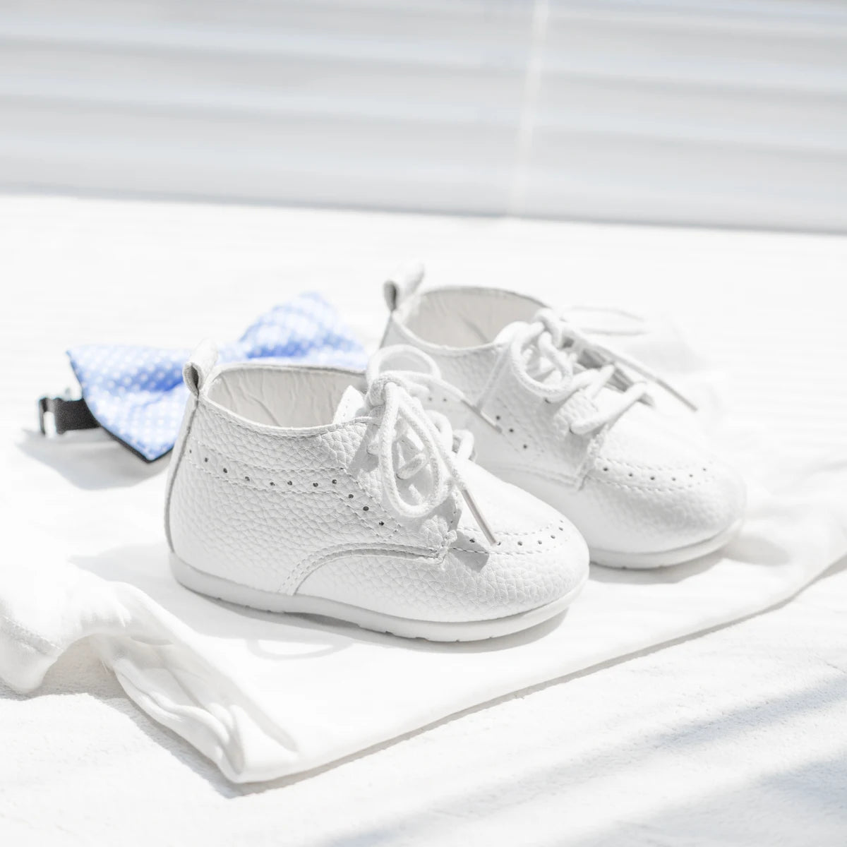 Flat lay of Kidsely white pebbled infant dress shoes styled next to a light blue formal baby bowtie.