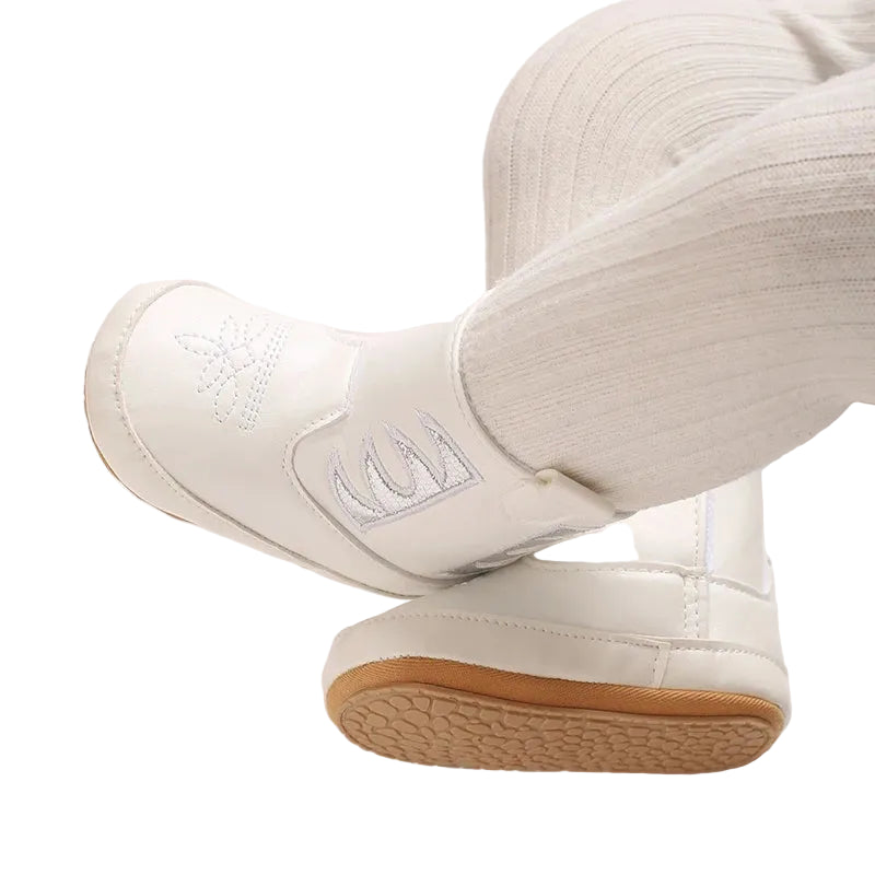 A baby wearing white and silver western cowboy booties with white tights, legs crossed showing the metallic side cutouts and toe design.