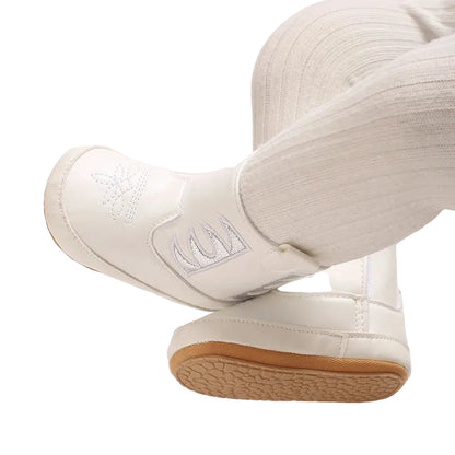 A baby wearing white and silver western cowboy booties with white tights, legs crossed showing the metallic side cutouts and toe design.
